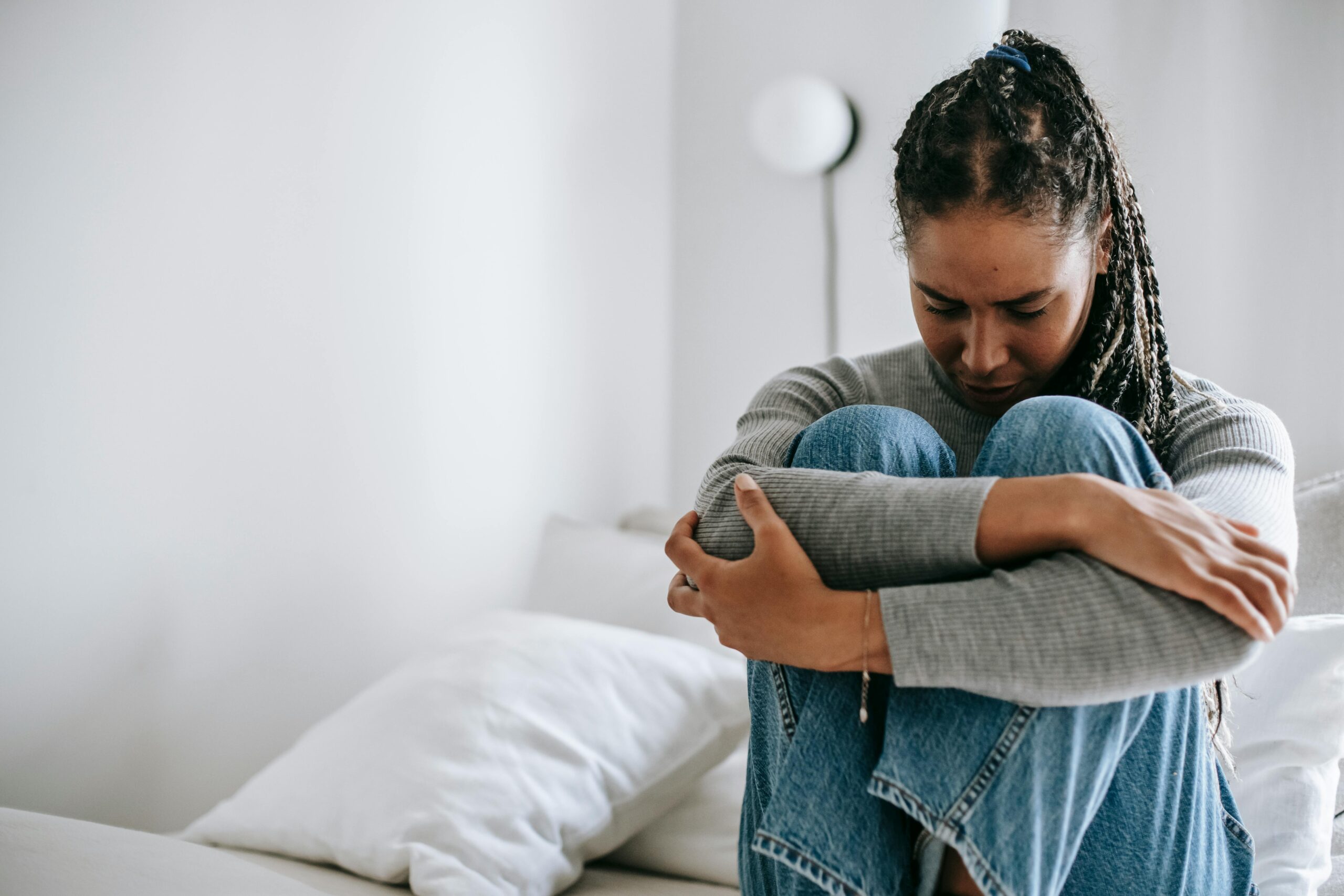 A young woman embraces her knees while sitting on a bed, appearing thoughtful and introspective.